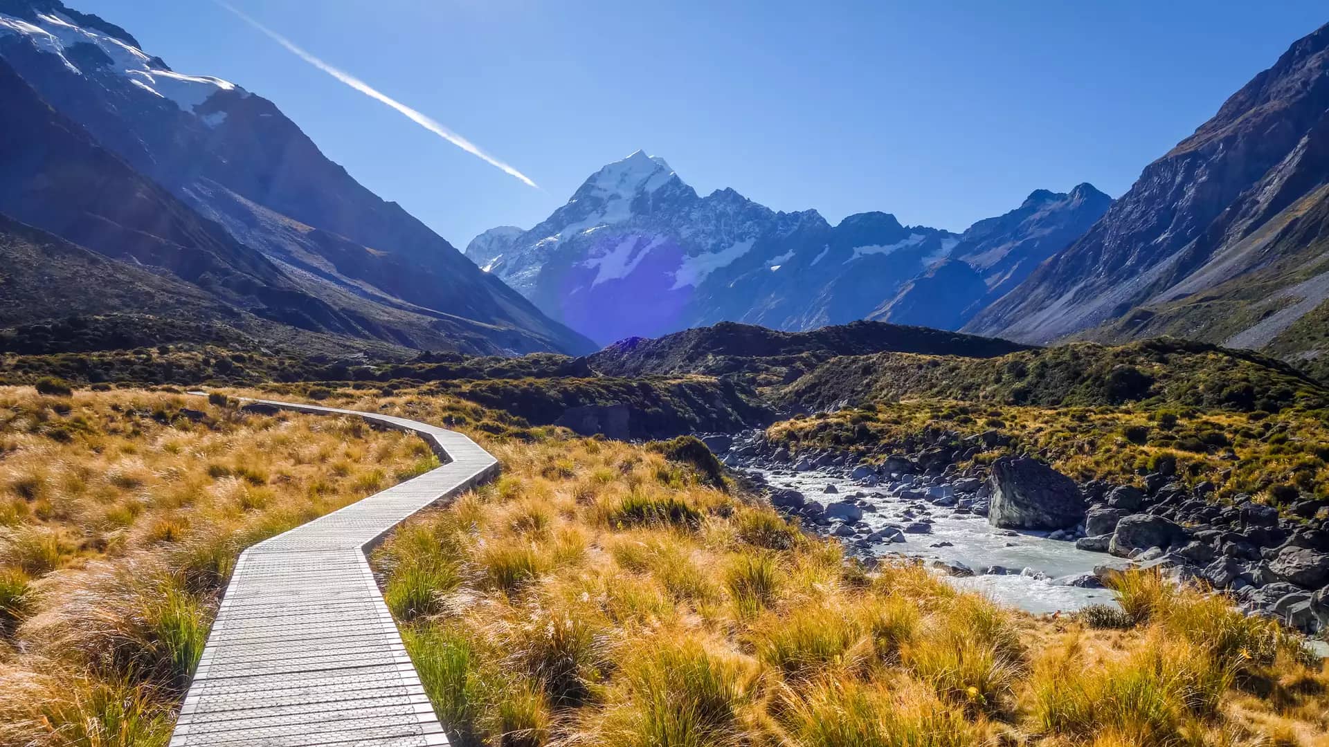 Boardwalk leading through grassland to snow-capped Aoraki Mount Cook under a bright blue sky. Credit: New Zealand Fine Touring Ltd
