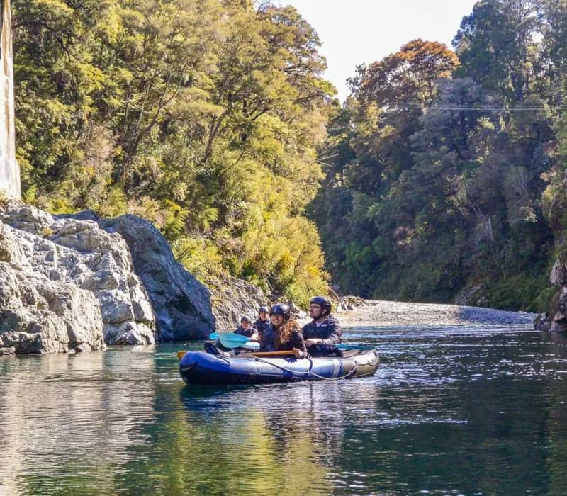 People kayaking on a calm river through a forested rocky landscape in bright daylight.