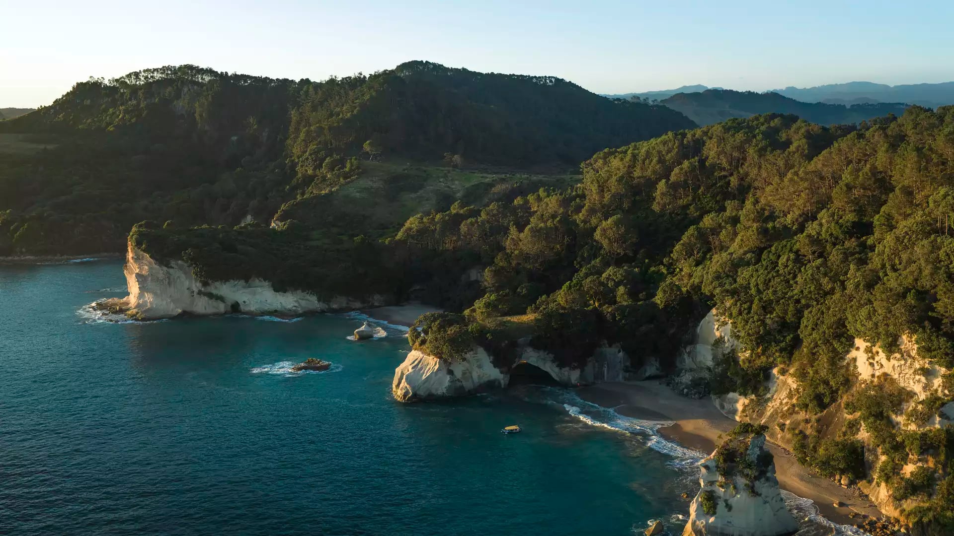 Aerial view of forested coastline, rocky cliffs, and beach with turquoise ocean water at sunset.