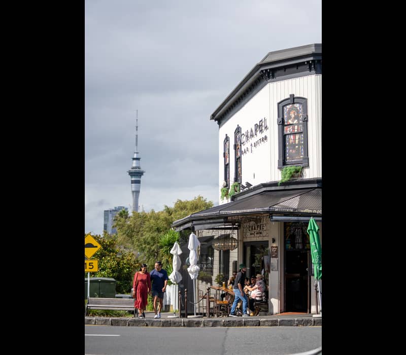 A couple walks along a sidewalk in Ponsonby, Auckland, passing the historic Chapel Bar & Bistro with its stained-glass windows, while the Sky Tower rises in the distant background under a cloudy sky. Credit: Tātaki Auckland Unlimited