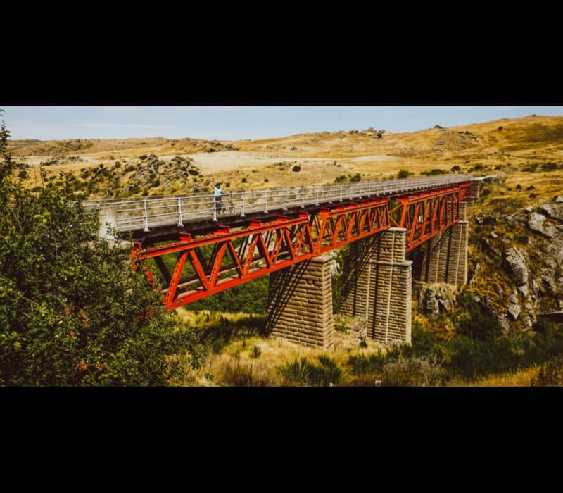 A cyclist stands on a high, red-steel lattice bridge supported by massive stone pillars, spanning a rugged rocky gorge in the dry, golden hills of Central Otago.