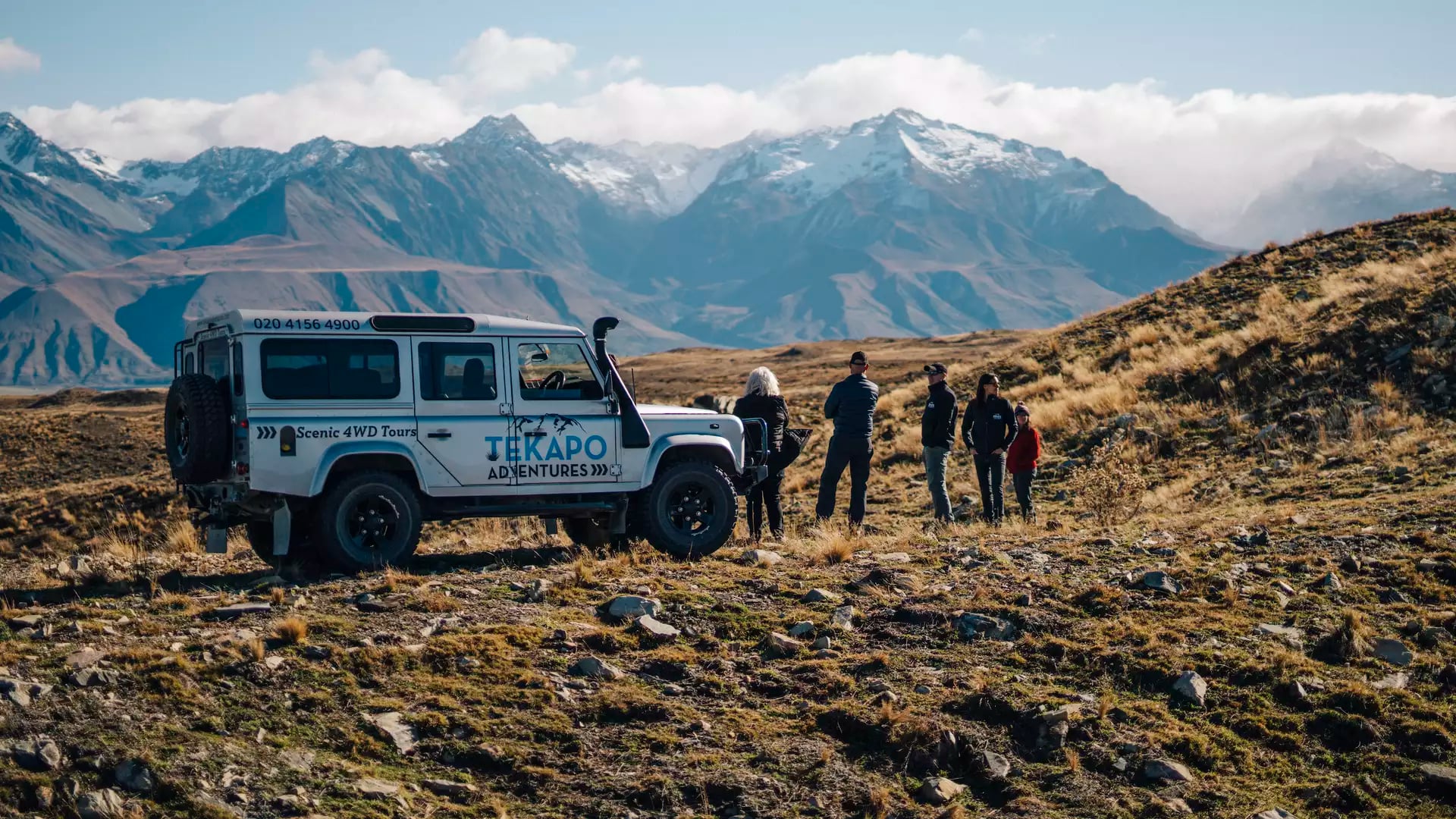 Small group stands by a 4WD vehicle with snow-capped mountains in the background during a tour.