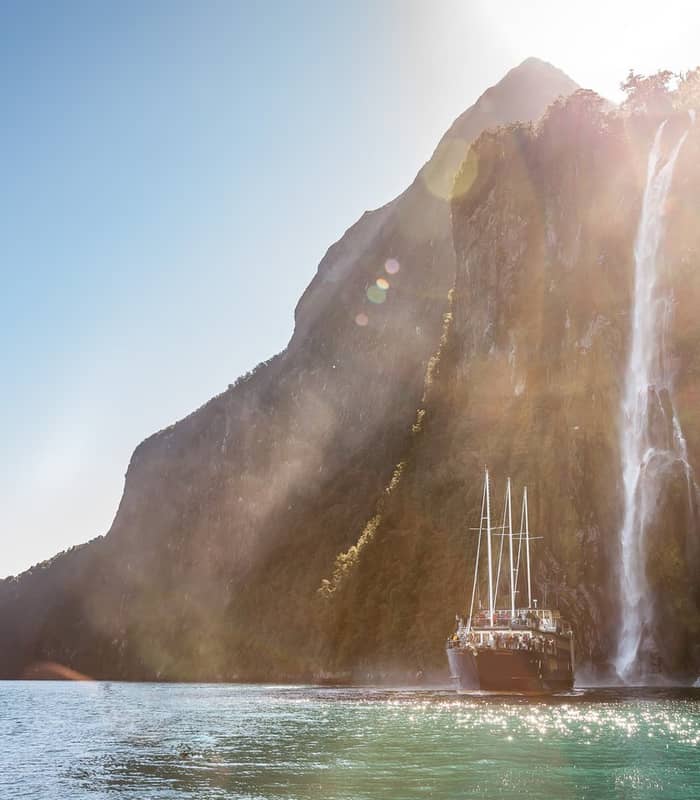 Tourist boat near a tall waterfall flowing down steep cliffs in Milford Sound with sunlight sparkling on the water.