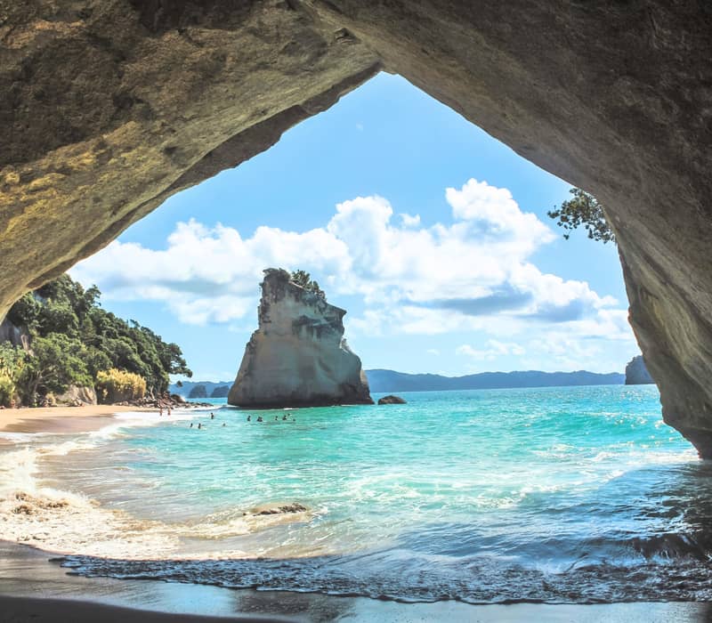 View from inside a cave of sandy beach, turquoise ocean, sea stack, and blue sky with clouds.