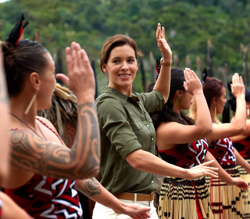 Group of women in traditional Maori attire performing a dance with raised hands outdoors.
