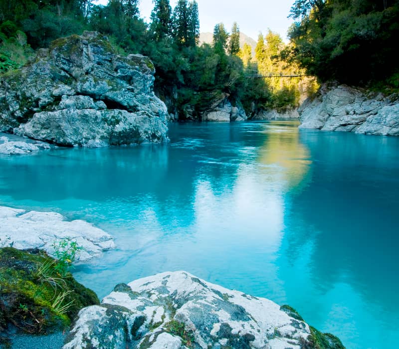 Blue river flowing between rocky banks and dense forest with sunlight reflecting on the water.