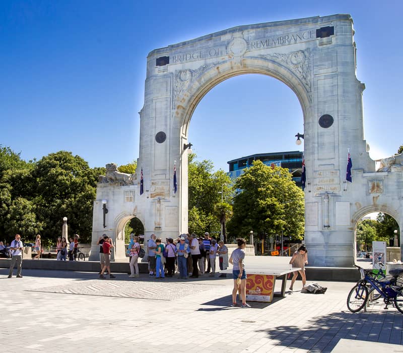 Stone memorial arch with tourists and bicycles under bright blue sky in a city plaza