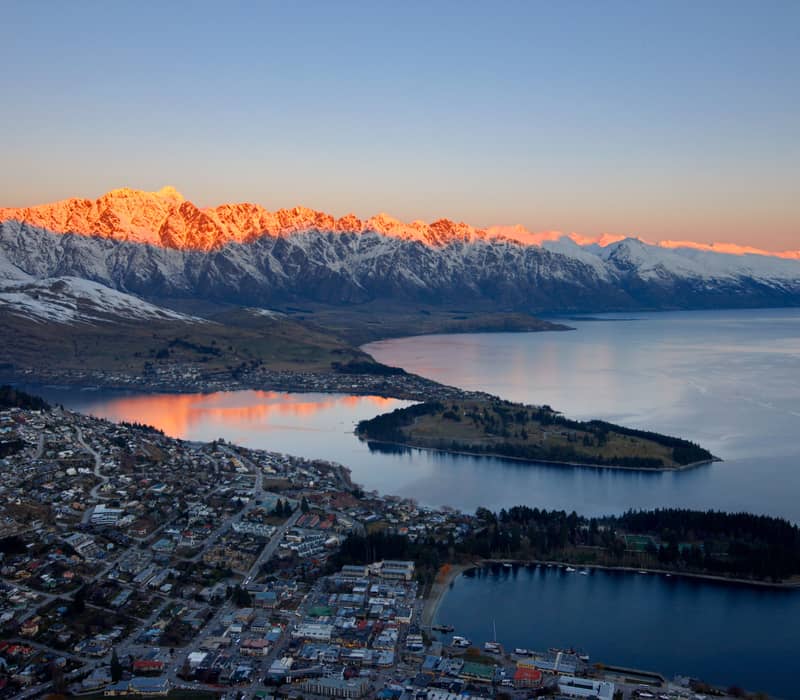 Sunset over Queenstown with pink snow-capped mountains, calm lake, and town buildings below.