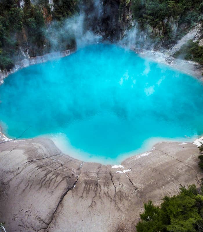 Bright blue geothermal crater lake with rising steam surrounded by green forest and rocky shore.
