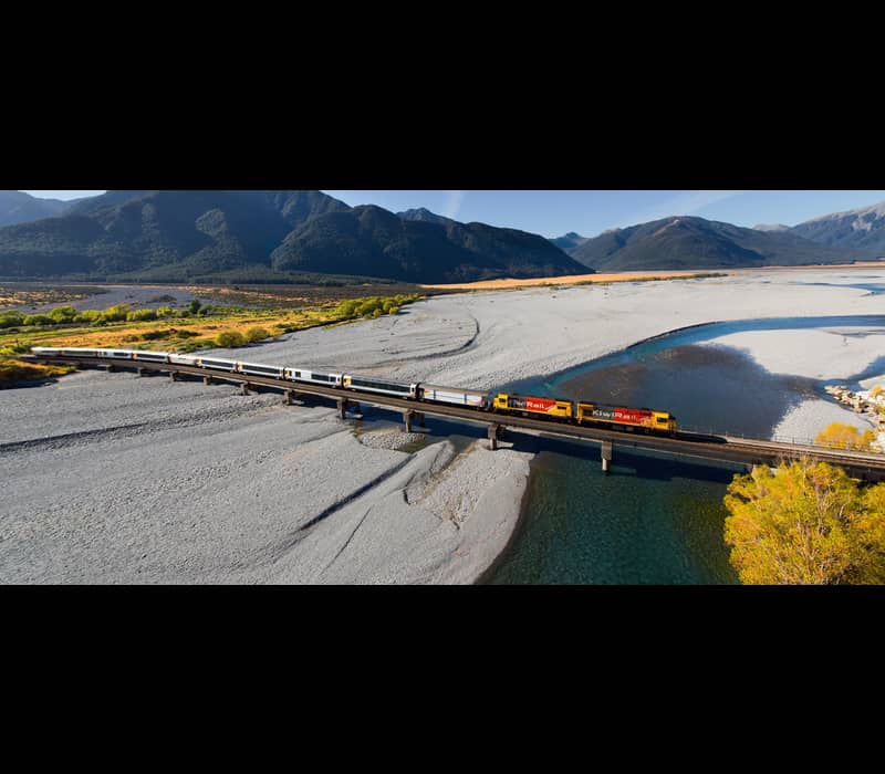 Train crossing bridge over river with mountains in background on a sunny day