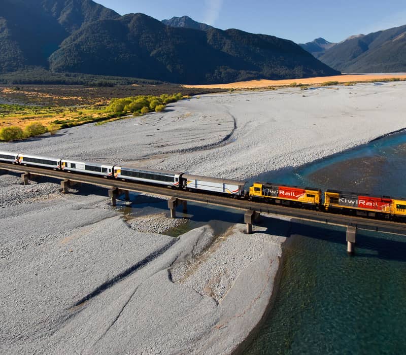 Train crossing bridge over river with mountains in background on a sunny day