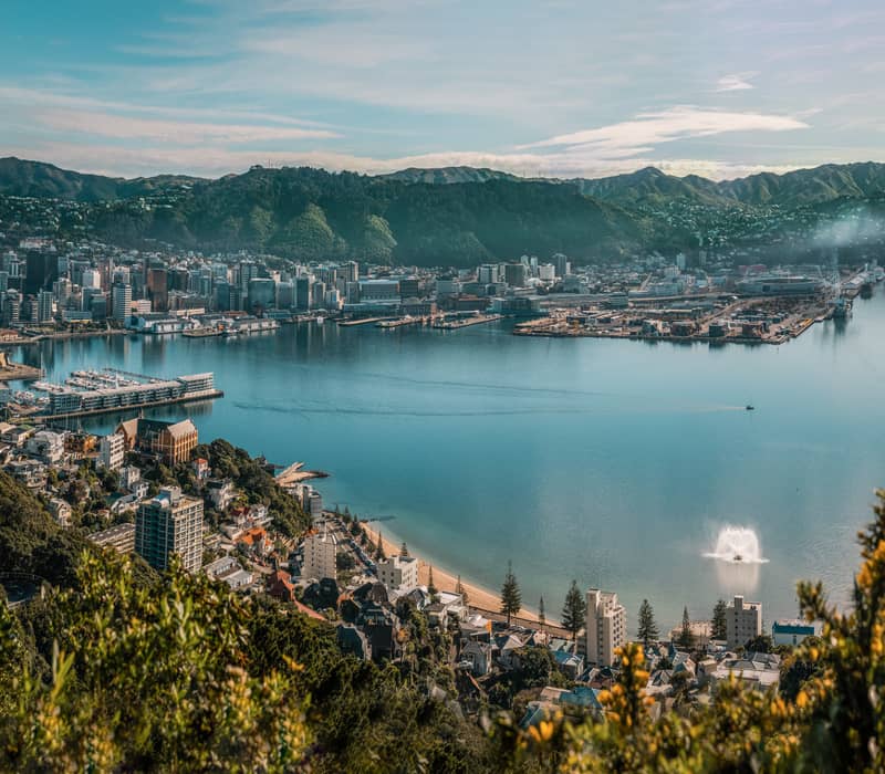 City view with harbor, waterfront buildings, and mountain backdrop under blue sky.