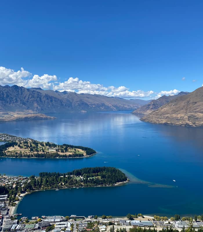 An expansive aerial view of Queenstown and Lake Wakatipu from a high vantage point, showing the Queenstown Gardens peninsula and the Remarkables mountain range under a clear blue sky.