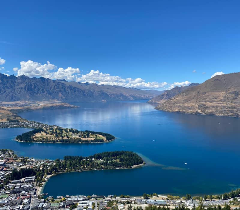 An expansive aerial view of Queenstown and Lake Wakatipu from a high vantage point, showing the Queenstown Gardens peninsula and the Remarkables mountain range under a clear blue sky.