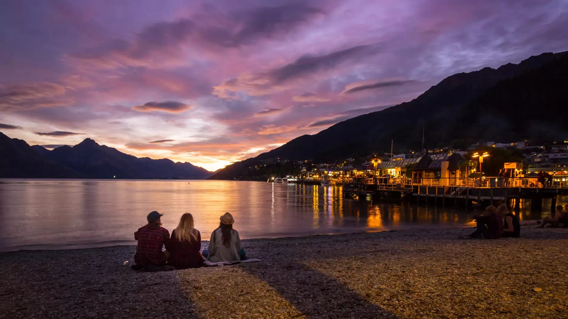 People sitting on a pebbled shore watching a colorful sunset over a lake with mountains and a lit dock.