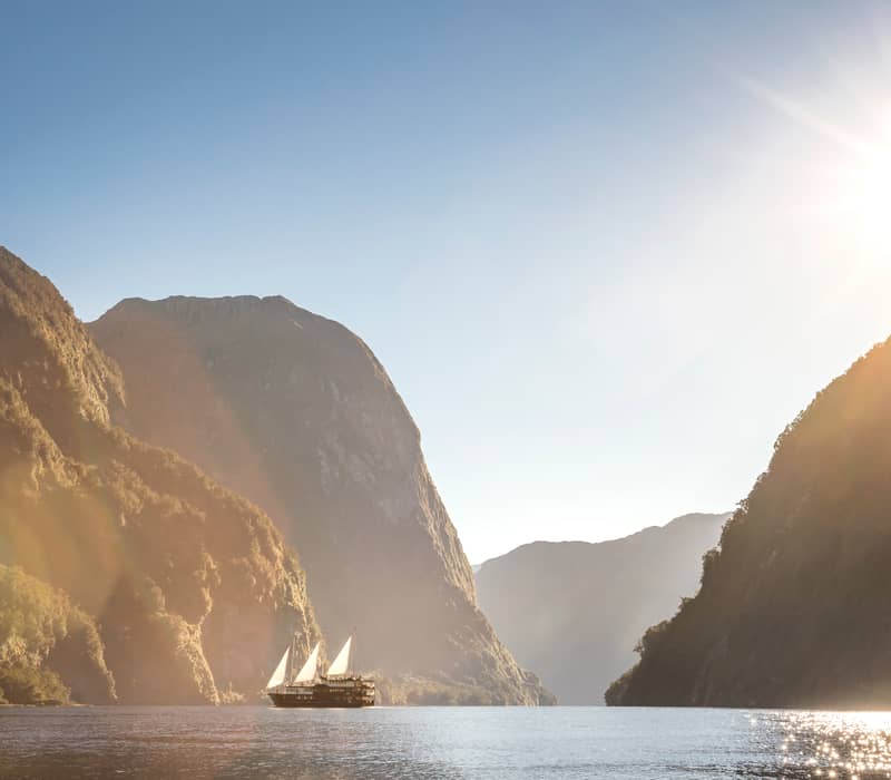 Sailboat sailing on calm fjord waters surrounded by tall mountains and sunlight.