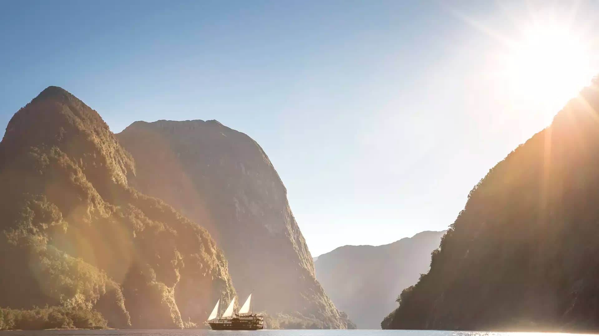 Sailboat sailing on calm fjord waters surrounded by tall mountains and sunlight.