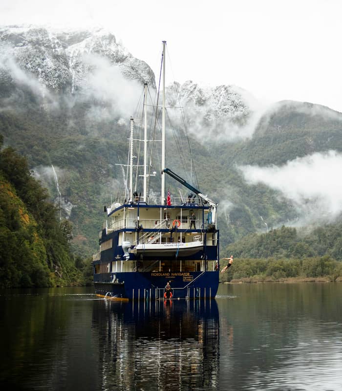 Large ship anchored in misty fjord with mountains and forests surrounding calm water.
