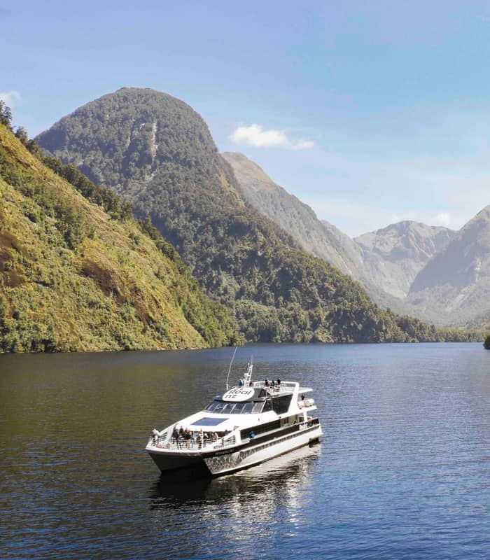 Boat sailing through calm fjord waters surrounded by green mountains under a clear sky.