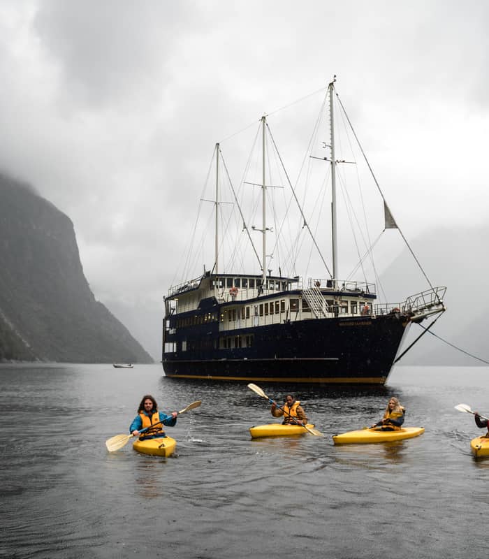 Four kayakers paddling in yellow kayaks near a cruise ship in a misty mountain fjord.