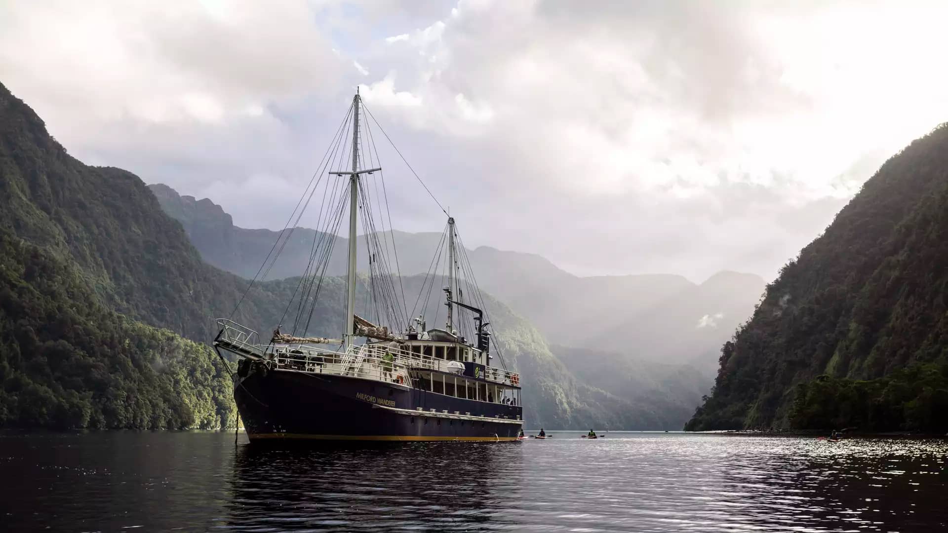 Boat on calm water between forest-covered mountains under cloudy sky with kayaks around