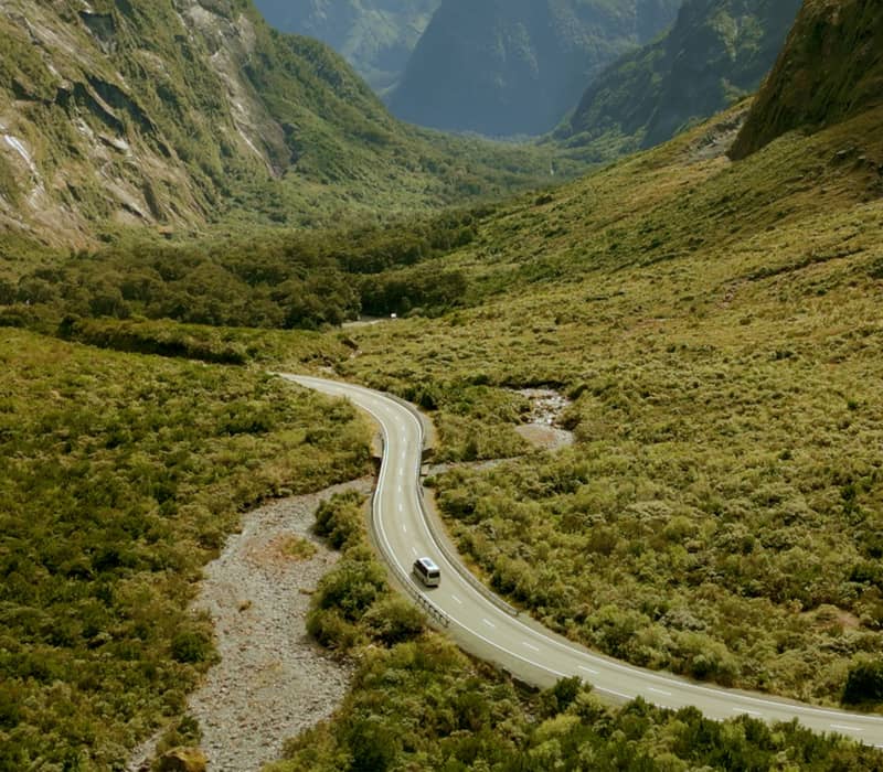 Van driving on winding road through green mountain valley with river nearby