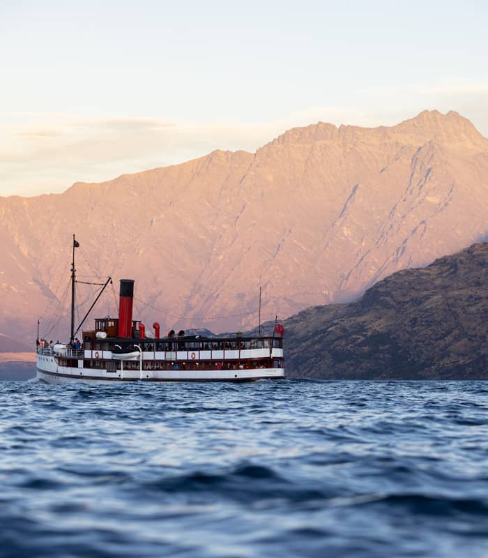 The historic TSS Earnslaw steamship cruises across the deep blue waters of Lake Wakatipu, framed by the golden peaks of the Remarkables at sunset.