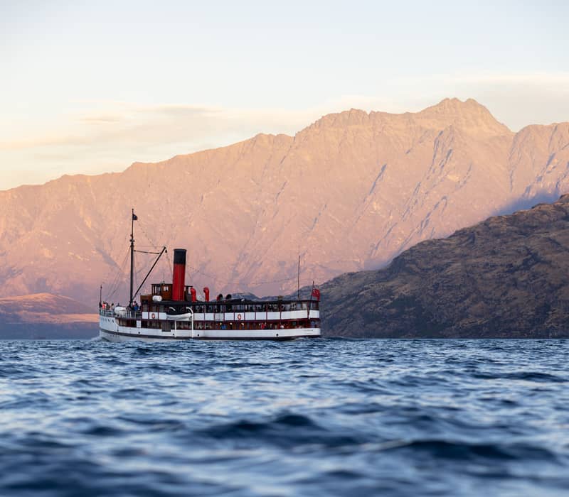 The historic TSS Earnslaw steamship cruises across the deep blue waters of Lake Wakatipu, framed by the golden peaks of the Remarkables at sunset.