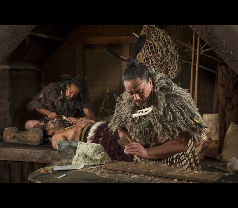 Maori men practicing traditional carving and tattooing with face tattoos and feathered clothing in a wooden workshop.
