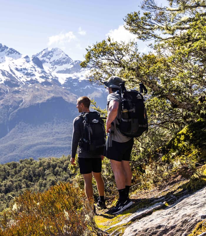 Two men with backpacks hiking on a trail with snowy mountains and forest in background.