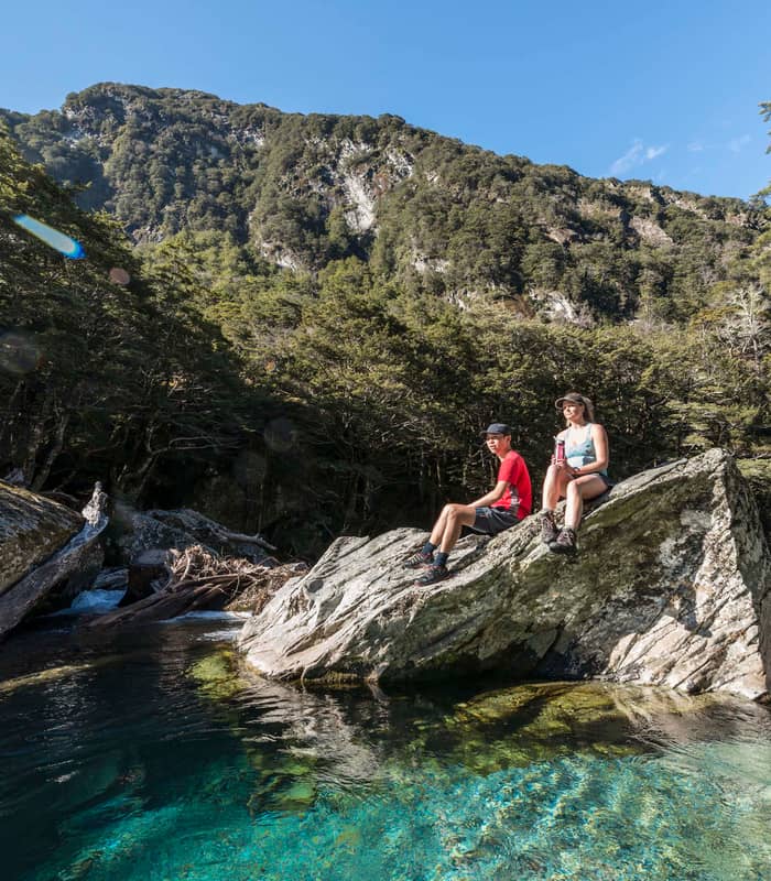 Two people sitting on rocks by a clear blue river with forest and mountains in background.