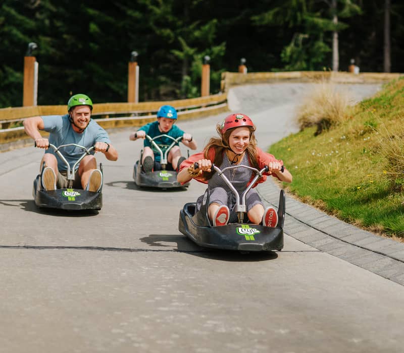 Three people race down the Skyline Queenstown Luge track in colorful carts, with Lake Wakatipu and the snow-capped Remarkables in the background.
