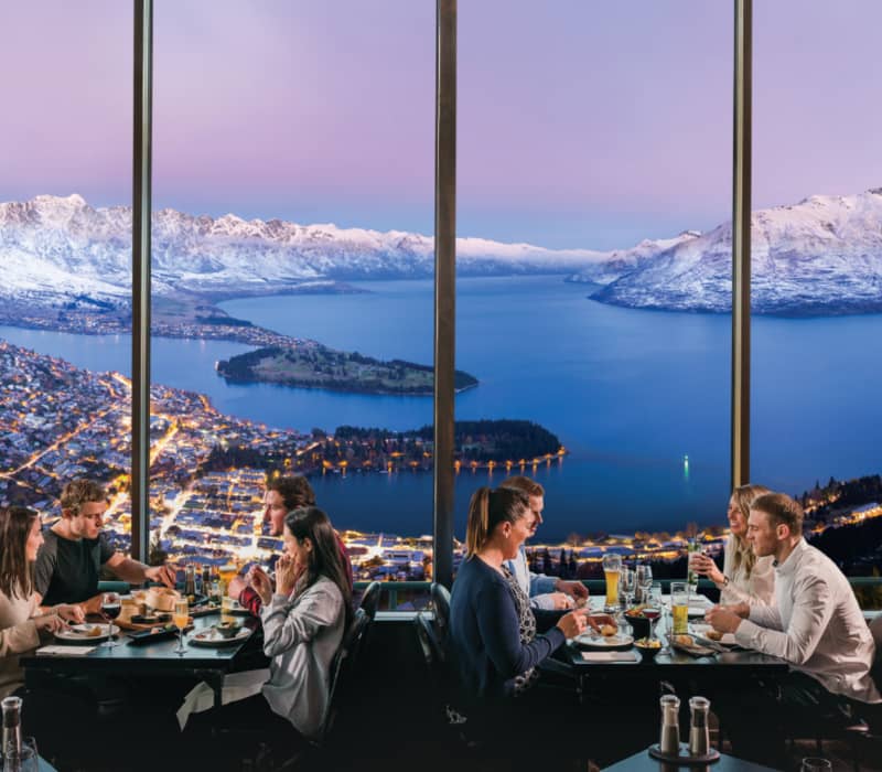 Groups of people dining at Stratosfare Restaurant at Skyline Queenstown, looking out through large windows at the city lights and snow-capped mountains at dusk.