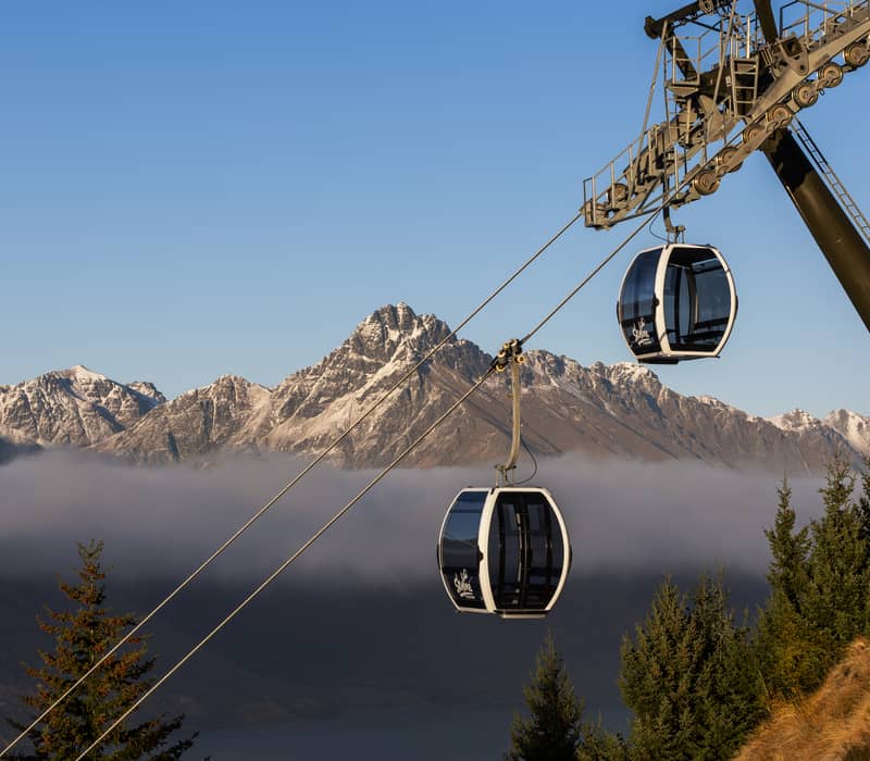 Two Skyline Queenstown gondola cabins travel along cables above a layer of low-lying clouds with the snow-dusted Remarkables in the background.