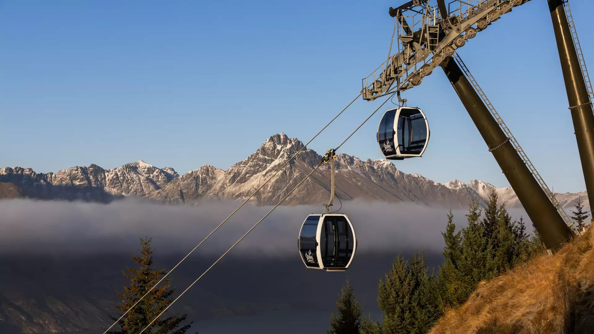 Two Skyline Queenstown gondola cabins travel along cables above a layer of low-lying clouds with the snow-dusted Remarkables in the background.