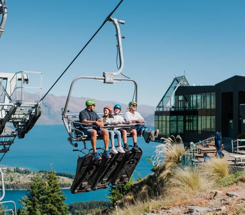 A family of four riding a chairlift with luge carts attached underneath, overlooking Lake Wakatipu at Skyline Queenstown.