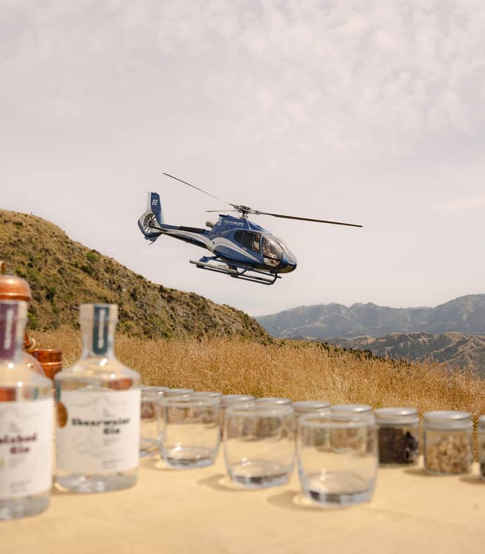 Helicopter flying over mountains with gin bottles and glasses on a table in the foreground.