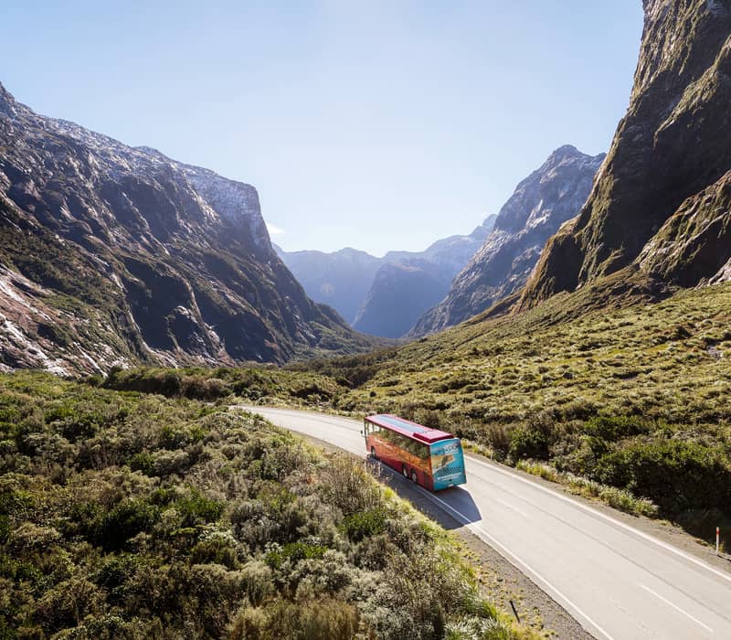 An elevated view of a red passenger coach driving along a winding road between steep forested mountain walls. Credit: Southern Discoveries