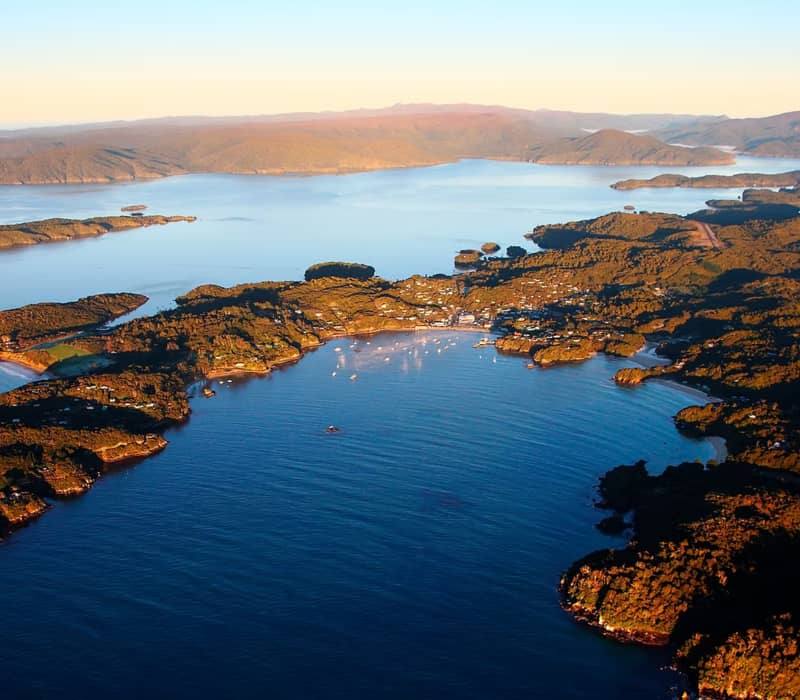An aerial view of Oban and Halfmoon Bay on Stewart Island during sunset, showing the township nestled around the coast with forested hills and Paterson Inlet in the distance.