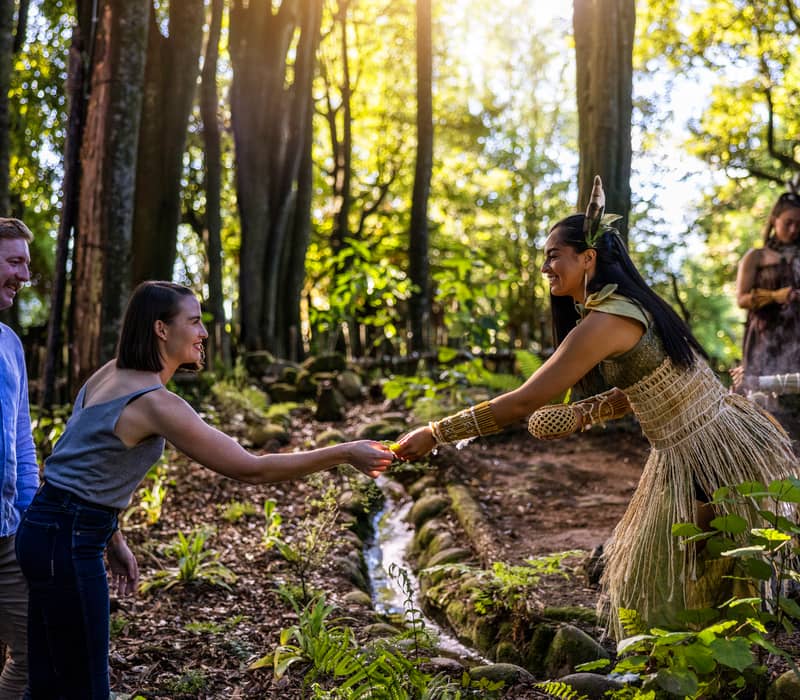 Tourists interacting with a woman in traditional dress in a sunlit forest.