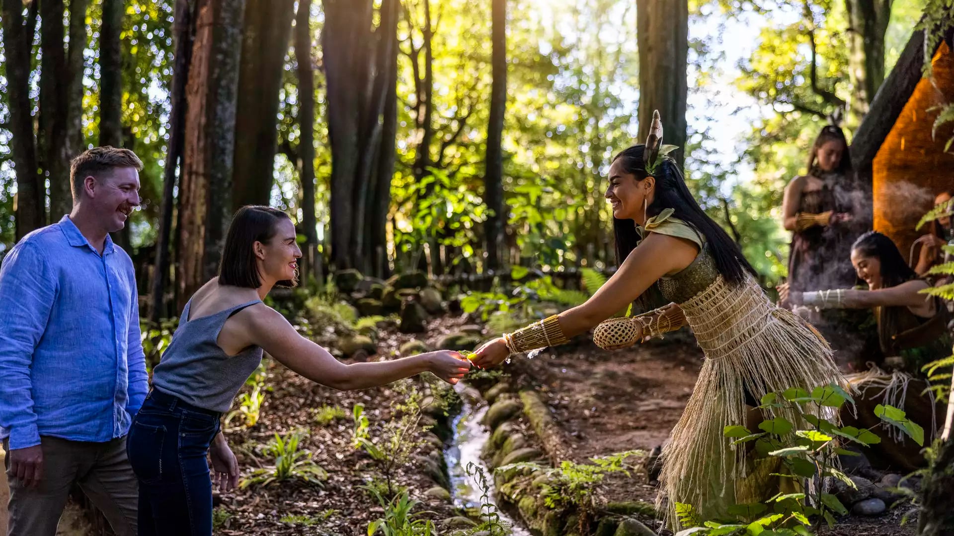 Tourists interacting with a woman in traditional dress in a sunlit forest.