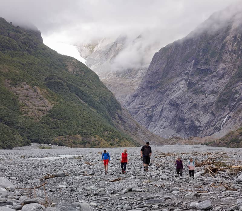 Five people hiking on rocky valley floor with mountains, clouds, and waterfalls in the background.