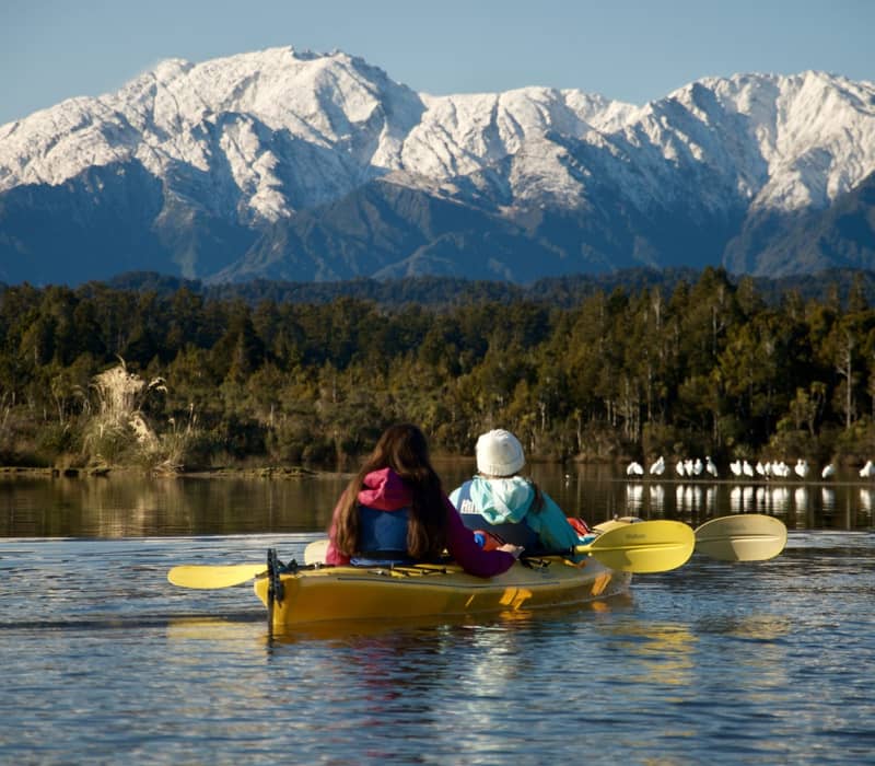 Two people in a yellow kayak on a lake with snowy mountains and forest in the background.