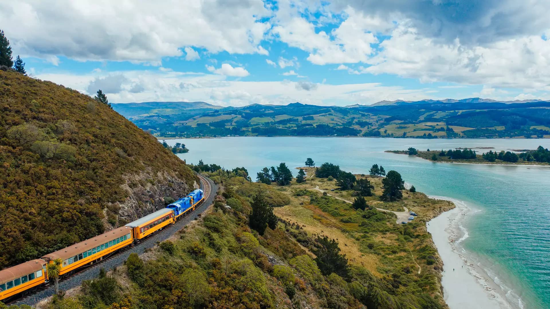 Train traveling through a green hillside next to a river and beach under a cloudy sky.