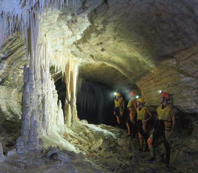 Four people with helmet lights explore an underground cave with stalactites and stalagmites.