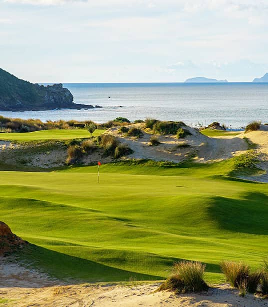 A world-class coastal golf course featuring bright green undulating fairways and a red flag, nestled among sandy dunes overlooking the blue Pacific Ocean under a soft sky.