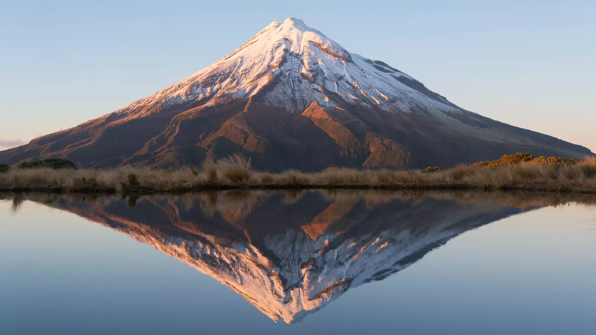 Snowcapped Mt Taranaki reflected clearly in a calm lake at sunset.