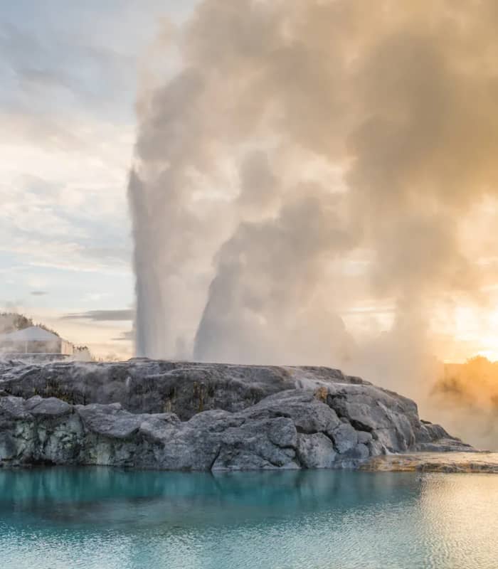 The Pōhutu Geyser erupts with a massive plume of steam and water behind a rocky silica terrace, reflected in a calm turquoise pool under a golden sky.