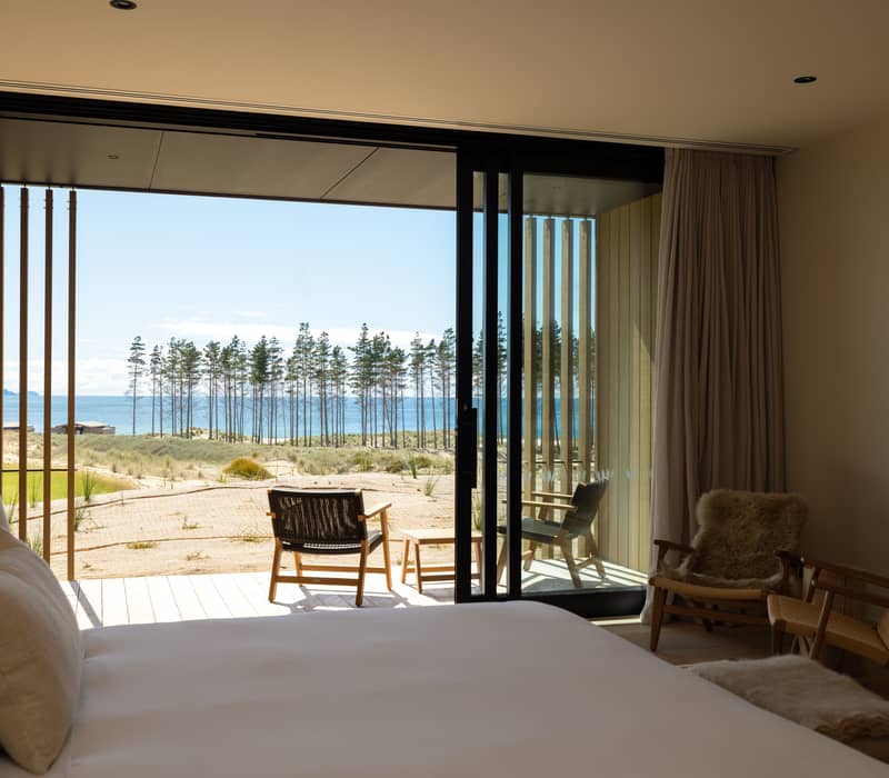 Bedroom with open sliding door showing balcony chairs and ocean with pine trees in the background.
