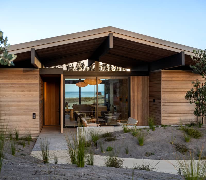 Modern wooden villa with glass doors overlooking beach, outdoor seating and sand dunes in front.